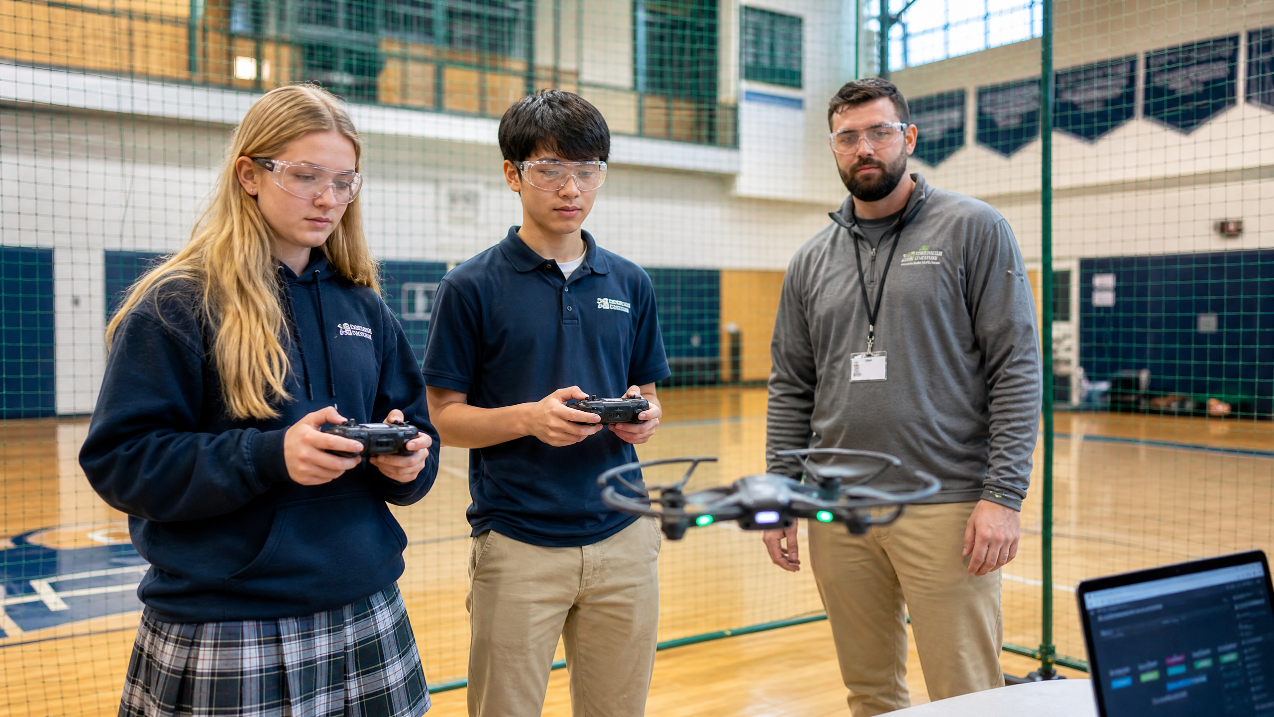 High school students wearing safety goggles flying indoor training drones inside a gymnasium safety netting enclosure